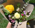Miniature bouquet of flowers in brown paper packaging held by a hand outdoors.